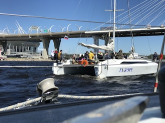 A group of people is on a sailboat named 'EUROPE' navigating under a large, modern bridge. The background includes a stadium and several structural elements of the bridge, with a bright blue sky overhead.