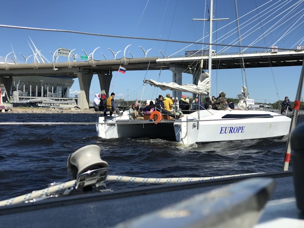 A group of people is on a sailboat named 'EUROPE' navigating under a large, modern bridge. The background includes a stadium and several structural elements of the bridge, with a bright blue sky overhead.