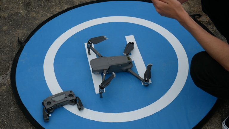 A drone rests on a circular blue landing pad with a white 'H' symbol. Next to it is a remote control, and a person's arm is visible holding something.