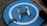 A drone rests on a circular blue landing pad with a white 'H' symbol. Next to it is a remote control, and a person's arm is visible holding something.