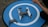 A drone rests on a circular blue landing pad with a white 'H' symbol. Next to it is a remote control, and a person's arm is visible holding something.