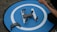 A drone rests on a circular blue landing pad with a white 'H' symbol. Next to it is a remote control, and a person's arm is visible holding something.