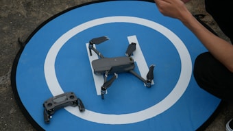 A drone rests on a circular blue landing pad with a white 'H' symbol. Next to it is a remote control, and a person's arm is visible holding something.