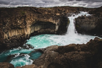 A powerful waterfall cascades into a vibrant turquoise pool surrounded by jagged rock formations. The sky overhead is filled with moody, dark clouds, adding a dramatic atmosphere to the scene. The rocks have interesting geological features, with columnar formations visible around the waterfall.