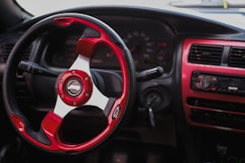 A car interior featuring a vibrant red and black sports steering wheel. The dashboard, including a speedometer and a radio unit, is visible with accents of red trim. The gear stick and air conditioning vents are also noticeable.