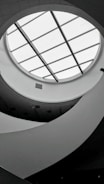 A monochromatic interior view featuring a circular skylight with grid-like glass panels. The ceiling design is modern, with curved, flowing architectural elements creating an abstract pattern. Soft lighting and shadows add depth to the scene.