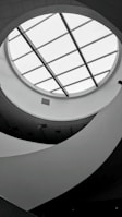 A monochromatic interior view featuring a circular skylight with grid-like glass panels. The ceiling design is modern, with curved, flowing architectural elements creating an abstract pattern. Soft lighting and shadows add depth to the scene.