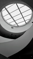 A monochromatic interior view featuring a circular skylight with grid-like glass panels. The ceiling design is modern, with curved, flowing architectural elements creating an abstract pattern. Soft lighting and shadows add depth to the scene.