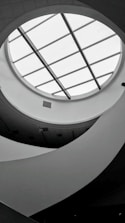 A monochromatic interior view featuring a circular skylight with grid-like glass panels. The ceiling design is modern, with curved, flowing architectural elements creating an abstract pattern. Soft lighting and shadows add depth to the scene.
