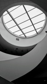 A monochromatic interior view featuring a circular skylight with grid-like glass panels. The ceiling design is modern, with curved, flowing architectural elements creating an abstract pattern. Soft lighting and shadows add depth to the scene.