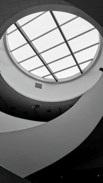A monochromatic interior view featuring a circular skylight with grid-like glass panels. The ceiling design is modern, with curved, flowing architectural elements creating an abstract pattern. Soft lighting and shadows add depth to the scene.