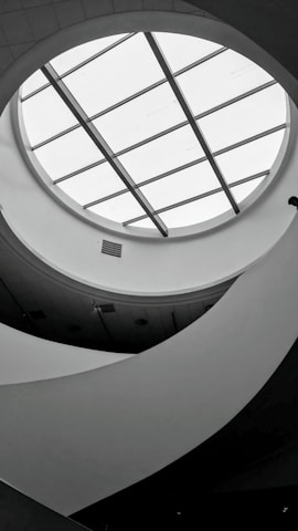 A monochromatic interior view featuring a circular skylight with grid-like glass panels. The ceiling design is modern, with curved, flowing architectural elements creating an abstract pattern. Soft lighting and shadows add depth to the scene.