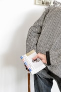 An elderly person holds a walking stick and a colorful pamphlet with their right hand. They are wearing a textured, patterned sweater over jeans. Part of a wall with a black-and-white photograph can be seen in the background.