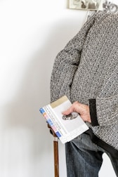 An elderly person holds a walking stick and a colorful pamphlet with their right hand. They are wearing a textured, patterned sweater over jeans. Part of a wall with a black-and-white photograph can be seen in the background.