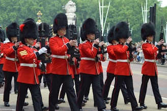 A group of soldiers in traditional ceremonial uniforms march in formation. They are wearing red coats and tall black bearskin hats, carrying rifles. The setting appears to be outdoors, with a road and trees in the background.