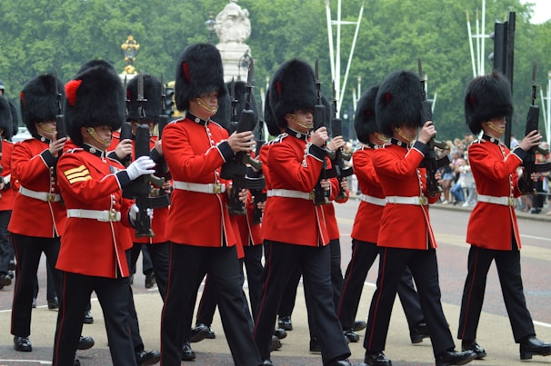 A group of soldiers in traditional ceremonial uniforms march in formation. They are wearing red coats and tall black bearskin hats, carrying rifles. The setting appears to be outdoors, with a road and trees in the background.