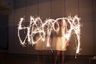 A person is playing with sparklers, creating bright, flowing trails of light against a dark, textured wall backdrop. The motion creates a dynamic and artistic light show.