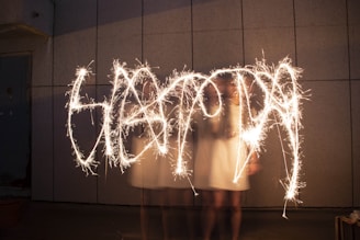A person is playing with sparklers, creating bright, flowing trails of light against a dark, textured wall backdrop. The motion creates a dynamic and artistic light show.