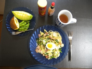 A tabletop setup featuring a meal with a plate of spaghetti garnished with sliced boiled egg, bacon, and vegetables. A smaller plate holds a sliced melon, edamame, and a pastry bun. Two bottles, possibly of sauce, and a mug of tea sit nearby, alongside a fork and knife.