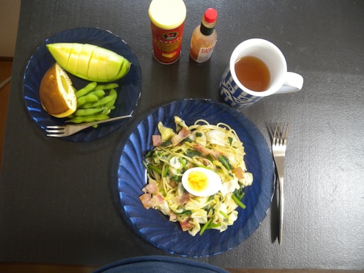 A tabletop setup featuring a meal with a plate of spaghetti garnished with sliced boiled egg, bacon, and vegetables. A smaller plate holds a sliced melon, edamame, and a pastry bun. Two bottles, possibly of sauce, and a mug of tea sit nearby, alongside a fork and knife.