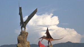 Two large sculptures of pterosaurs standing on rock formations against a backdrop of a blue sky with some clouds. The pterosaurs appear in dynamic poses, with their wings spread, adding to the dramatic effect of the scene.