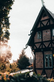 A picturesque scene featuring a quaint half-timbered house with intricate brickwork and dark wooden beams. The gable roof is steeply pitched and adorned with decorative elements. Sunlight filters through the surrounding trees, casting warm, dappled light on the building. The foreground features lush greenery, adding to the serene atmosphere.
