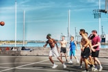 A group of men are playing basketball on an outdoor court near a waterfront area. The sky is clear and blue, and there are cranes and industrial structures visible in the background. The players are actively engaged, with one person shooting the ball while the others look on. The court has a modern design, and there is sports equipment laying on a bench nearby.