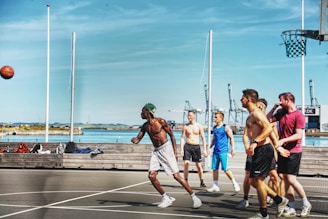 A group of men are playing basketball on an outdoor court near a waterfront area. The sky is clear and blue, and there are cranes and industrial structures visible in the background. The players are actively engaged, with one person shooting the ball while the others look on. The court has a modern design, and there is sports equipment laying on a bench nearby.