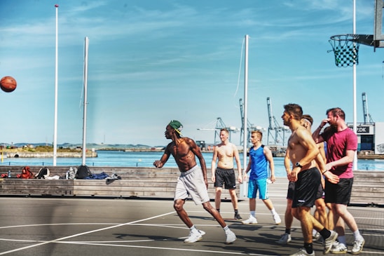 A group of men are playing basketball on an outdoor court near a waterfront area. The sky is clear and blue, and there are cranes and industrial structures visible in the background. The players are actively engaged, with one person shooting the ball while the others look on. The court has a modern design, and there is sports equipment laying on a bench nearby.