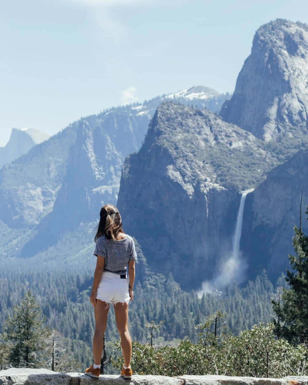 woman wearing blue shirt and white shorts standing beside rocky mountain during daytime