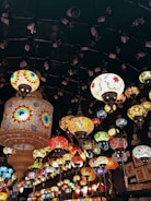 A collection of vibrant, intricately designed mosaic lamps hang from the ceiling, creating a colorful tapestry of light. The lamps vary in shape and size, featuring geometric patterns and a rich palette of colors. The atmosphere is warm and inviting, with a bohemian or traditional Middle Eastern aesthetic.