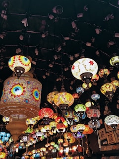 A collection of vibrant, intricately designed mosaic lamps hang from the ceiling, creating a colorful tapestry of light. The lamps vary in shape and size, featuring geometric patterns and a rich palette of colors. The atmosphere is warm and inviting, with a bohemian or traditional Middle Eastern aesthetic.