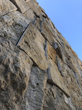 A close-up perspective of a stone wall composed of roughly hewn, large rectangular blocks stacked atop one another, displaying their natural rugged texture and grooves. The blue sky is visible in the background, creating a stark contrast with the earthy tones of the stones.