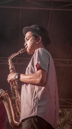 A musician wearing a hat and a patterned short-sleeve shirt plays the saxophone. The saxophonist is captured in profile, concentrating on their performance. The setting suggests an outdoor event with a stage background. There's a sense of focus and dedication in the musician's demeanor.