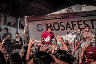 A lively music concert with a band performing on stage. The crowd is engaged, raising their hands in enthusiasm, and the backdrop reads 'MOSAFEST' indicating a festival setting.