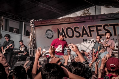 A lively music concert with a band performing on stage. The crowd is engaged, raising their hands in enthusiasm, and the backdrop reads 'MOSAFEST' indicating a festival setting.
