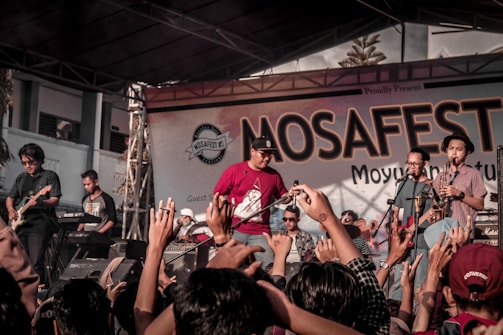 A lively music concert with a band performing on stage. The crowd is engaged, raising their hands in enthusiasm, and the backdrop reads 'MOSAFEST' indicating a festival setting.