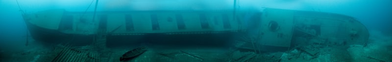 The side of a sunken ship resting in murky green water, with broken timbers scattered across the silty bottom