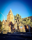 A picturesque stone church with a tall, pointed tower and arched windows, surrounded by palm trees. The building showcases a mix of stonework and red brick detailing, with artistic windows and a decorative entrance. Sunlight casts sharp shadows on the church and the surrounding area, creating a dramatic visual effect.