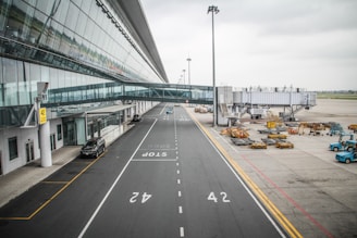 A modern airport terminal with a glass facade and an elevated walkway leading to a jet bridge. The tarmac is visible with service vehicles, parked luggage carts, and equipment scattered around. Sparse clouds fill the sky, creating a calm and orderly atmosphere.