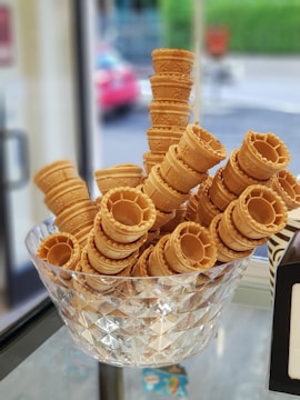 A large glass bowl containing multiple stacks of waffle-style ice cream cones. The cones are arranged in a way that forms various heights within the bowl. The background features a blurred view of the outside street, with some greenery and a hint of a red car.