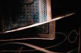A close-up shot of a playing card with intricate patterns in dark and moody lighting. The card's design includes geometric and floral elements, and it partially rests inside a reflective glass. Soft light creates highlights and shadows, accentuating the textures.