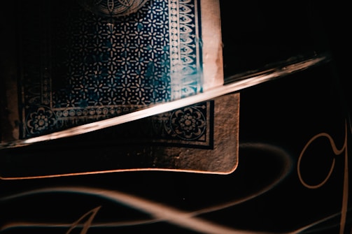 A close-up shot of a playing card with intricate patterns in dark and moody lighting. The card's design includes geometric and floral elements, and it partially rests inside a reflective glass. Soft light creates highlights and shadows, accentuating the textures.