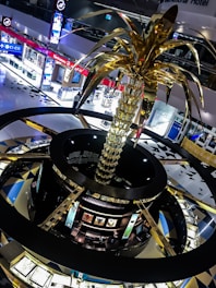 A large, ornate sculpture resembling a palm tree with golden fronds dominates the interior of a shopping mall. The mall features brightly lit electronic stores and jeweler displays, creating a modern and luxurious atmosphere. The architecture includes circular design elements with contrasting dark and gold colors.