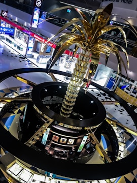 A large, ornate sculpture resembling a palm tree with golden fronds dominates the interior of a shopping mall. The mall features brightly lit electronic stores and jeweler displays, creating a modern and luxurious atmosphere. The architecture includes circular design elements with contrasting dark and gold colors.