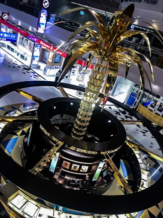 A large, ornate sculpture resembling a palm tree with golden fronds dominates the interior of a shopping mall. The mall features brightly lit electronic stores and jeweler displays, creating a modern and luxurious atmosphere. The architecture includes circular design elements with contrasting dark and gold colors.
