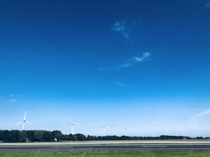 A landscape features several wind turbines aligned along a horizon with a backdrop of dense green trees. The sky above is expansive and vividly blue, with a few scattered clouds. In the foreground, there is a strip of grass adjacent to a highway or road.