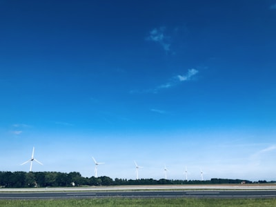 A landscape features several wind turbines aligned along a horizon with a backdrop of dense green trees. The sky above is expansive and vividly blue, with a few scattered clouds. In the foreground, there is a strip of grass adjacent to a highway or road.