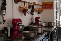 A kitchen setup with various cooking appliances and utensils neatly arranged. There are red stand mixers on the counter alongside several stainless steel pots and pans. Above the counter, copper utensils and scoops are hung on a rack. Bottles of olive oil and black pepper are also visible. The environment looks organized and ready for cooking.
