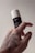 A hand with a tattooed design holds a cylindrical container with a black label and white cap. The label reads 'KORRES', 'Pinho Negro', and 'Black Pine'. The background is a neutral, soft color, highlighting the product and the hand.